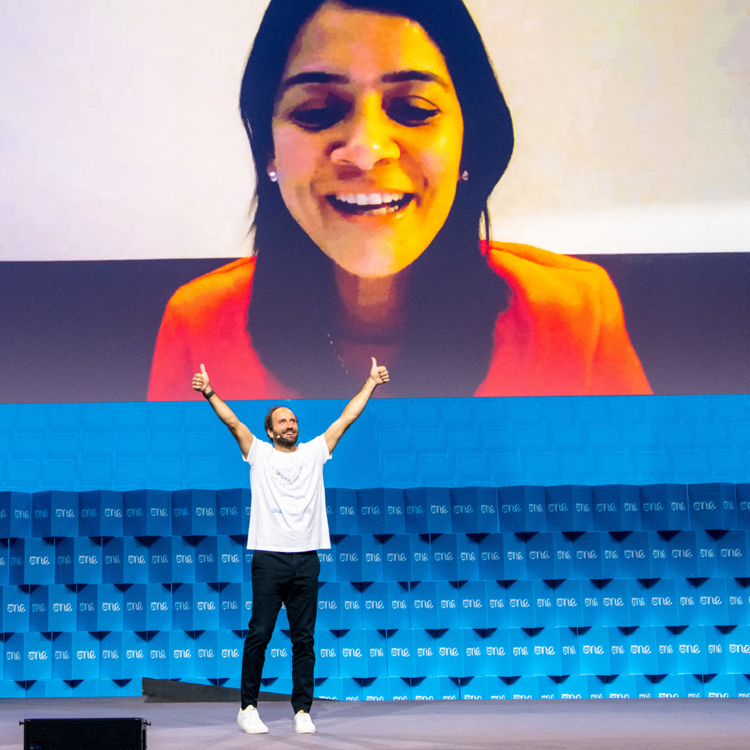 A man wearing a white t-shirt stands on a stage. His arms reach up towards a large screen projecting a picture of a woman. She is looking down at him.