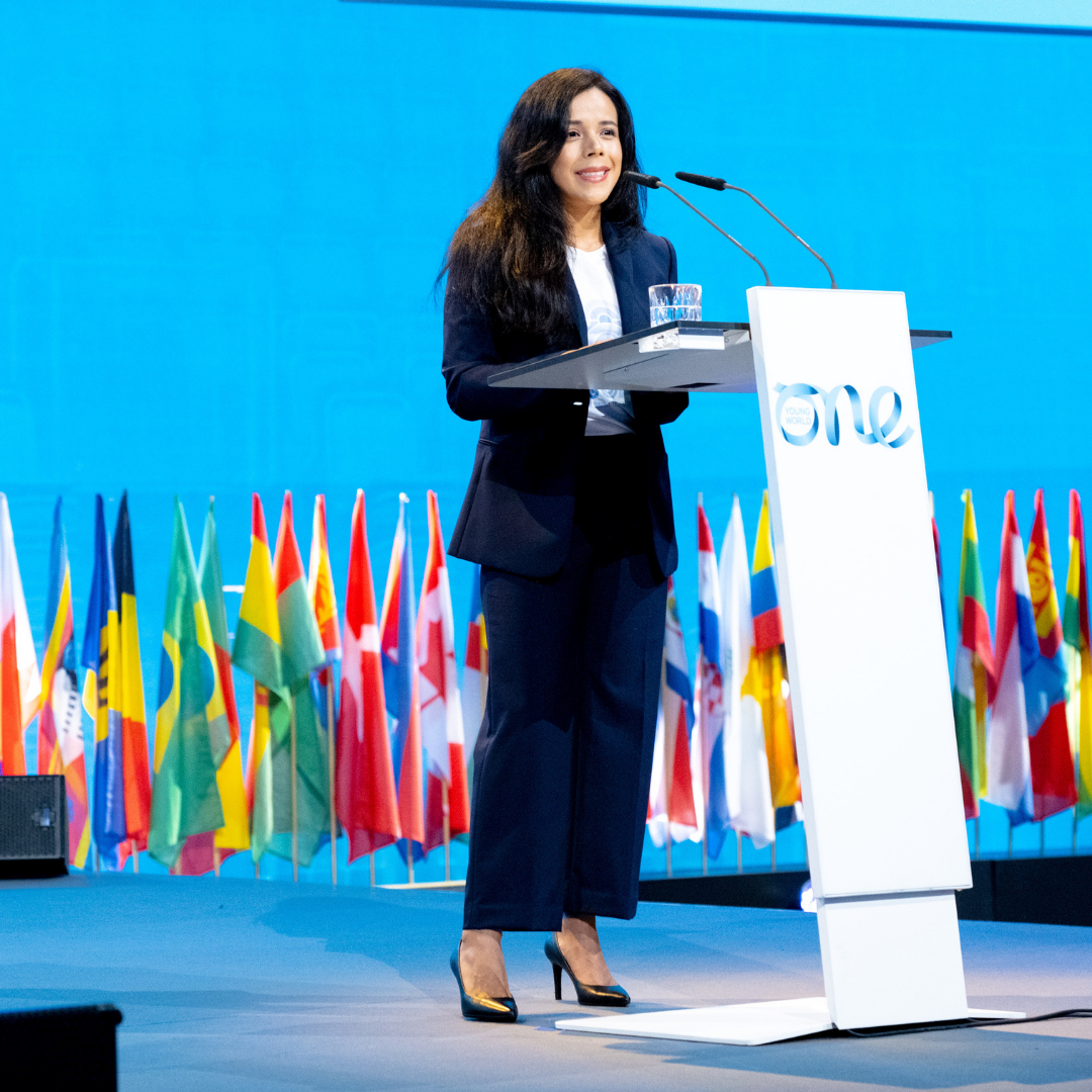 A young women in a dark suit stands at a white podium with the One Young World logo shown on the front. She smiles as she appears to speak to a crowd.