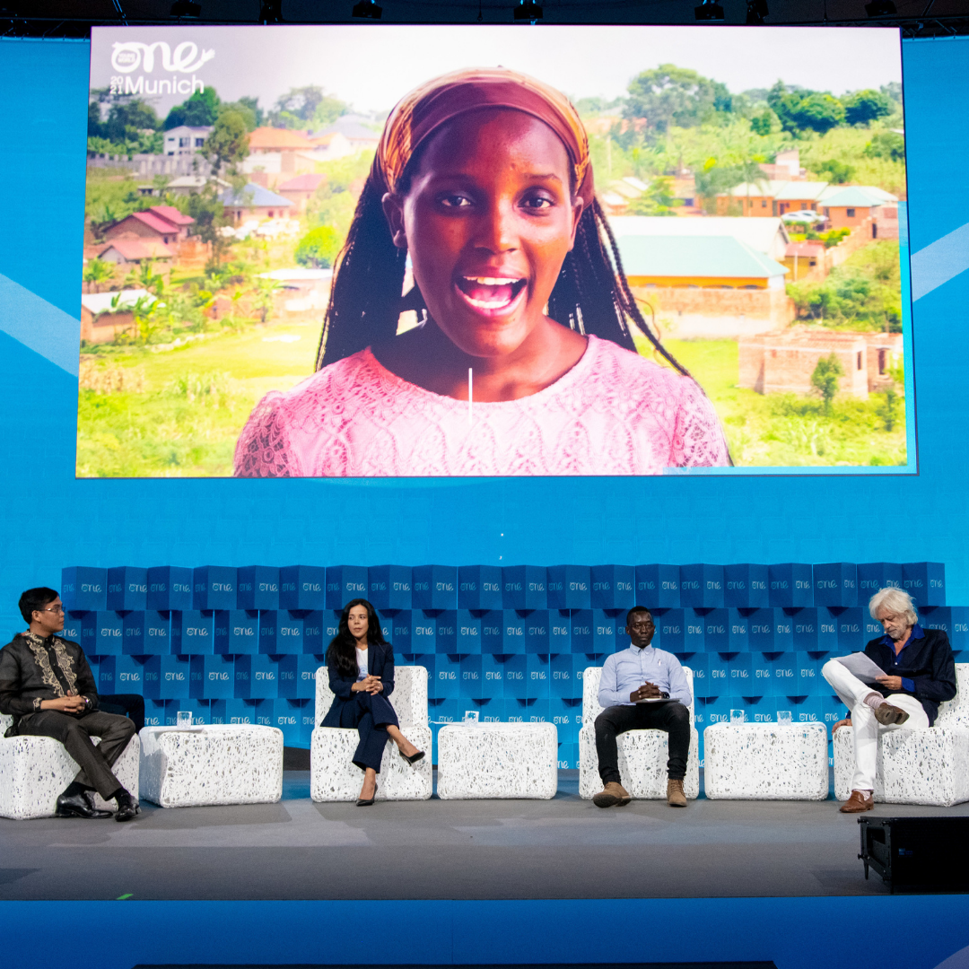 A young women speaks from a screen projected above a stage. Four people sit under the screen, listening. They sit on white chairs against a blue backdrop with the One Young World logo repeated.