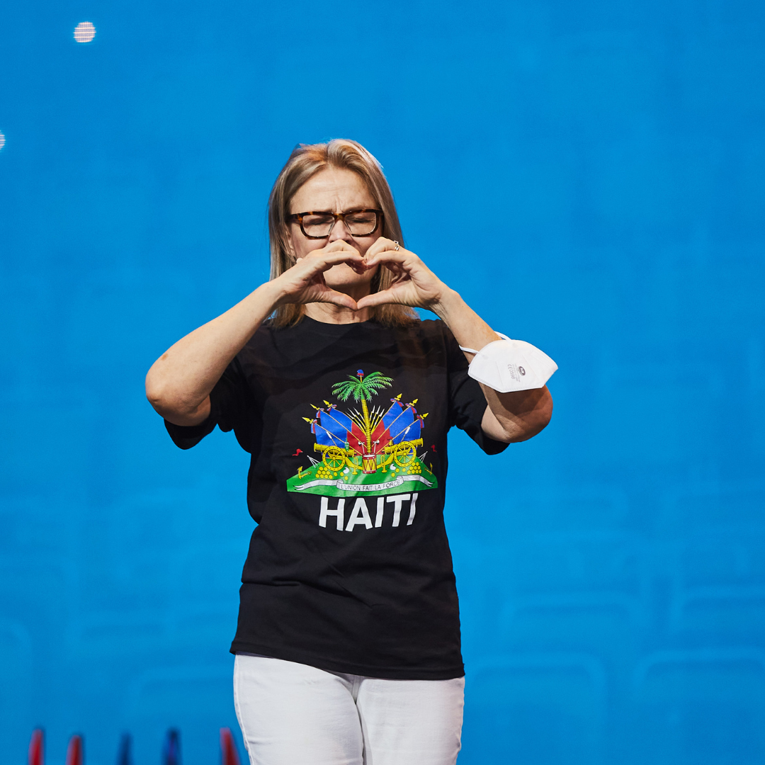 A woman [Kate Robertson] stands on stage looking at the camera. She creates a heart shape using both her hands. She is wearing a black t-shirt with a tropical design featuring the word 'HAITI' in capital letters.