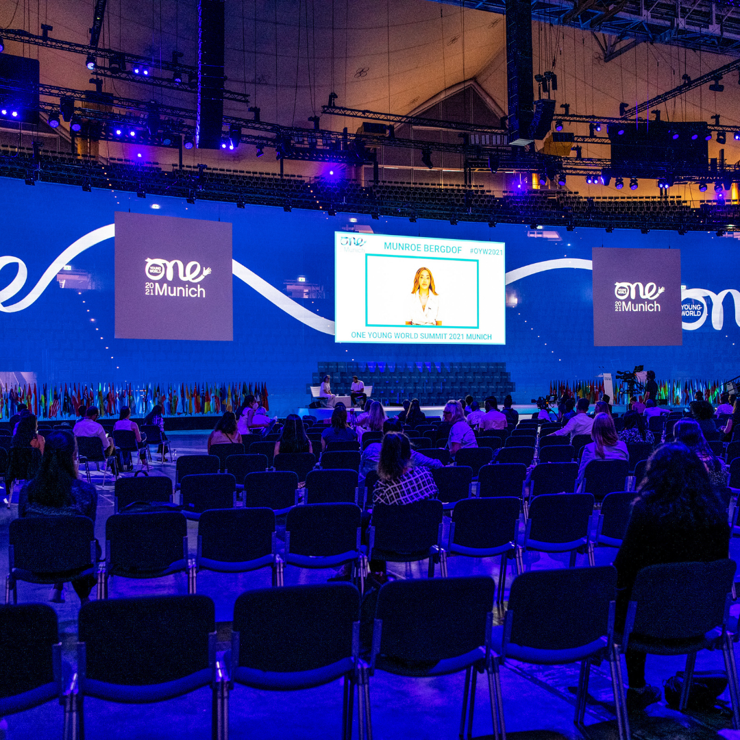 A wide angle image of a crowd sat in deep blue lighting. They are watching a screen projected onto stage. On screen is a woman [Munroe Bergdorf] wearing white, looking and speaking directly to the camera.