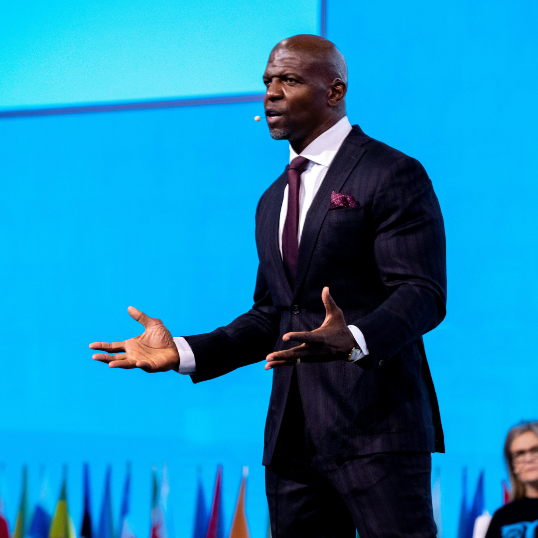 Terry Crews, Actor & One Young World Counsellor, speaks on stage with a blue backdrop. He wears a dark suit, standing with his arms outstretched as he addresses the crowd.