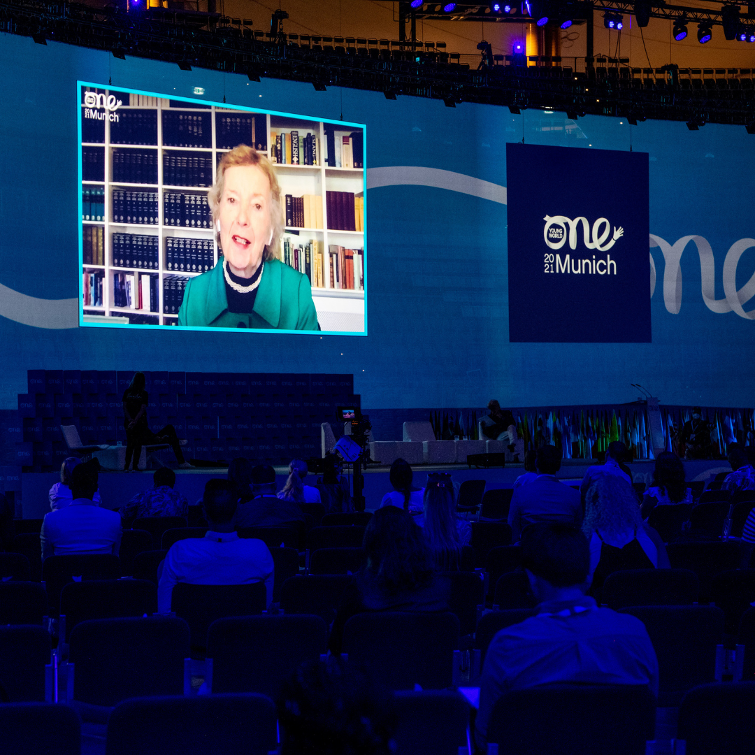 A crowd bathed in deep blue light watch a large screen projecting Mary Robinson, Former Prime Minister of Ireland, deliver a virtual keynote speech.