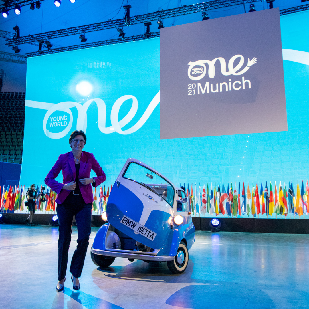 A women wearing a pink jacket and black trouser steps of out an old-style BMW vehicle. She is smiling and walking towards the camera.