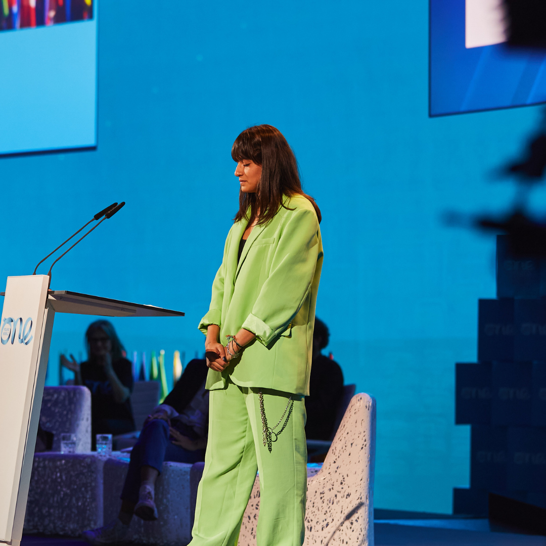 A women wearing a lime green suit stands on a stage against a blue backdrop. She is positioned behind a white podium with her head down and eyes closed.