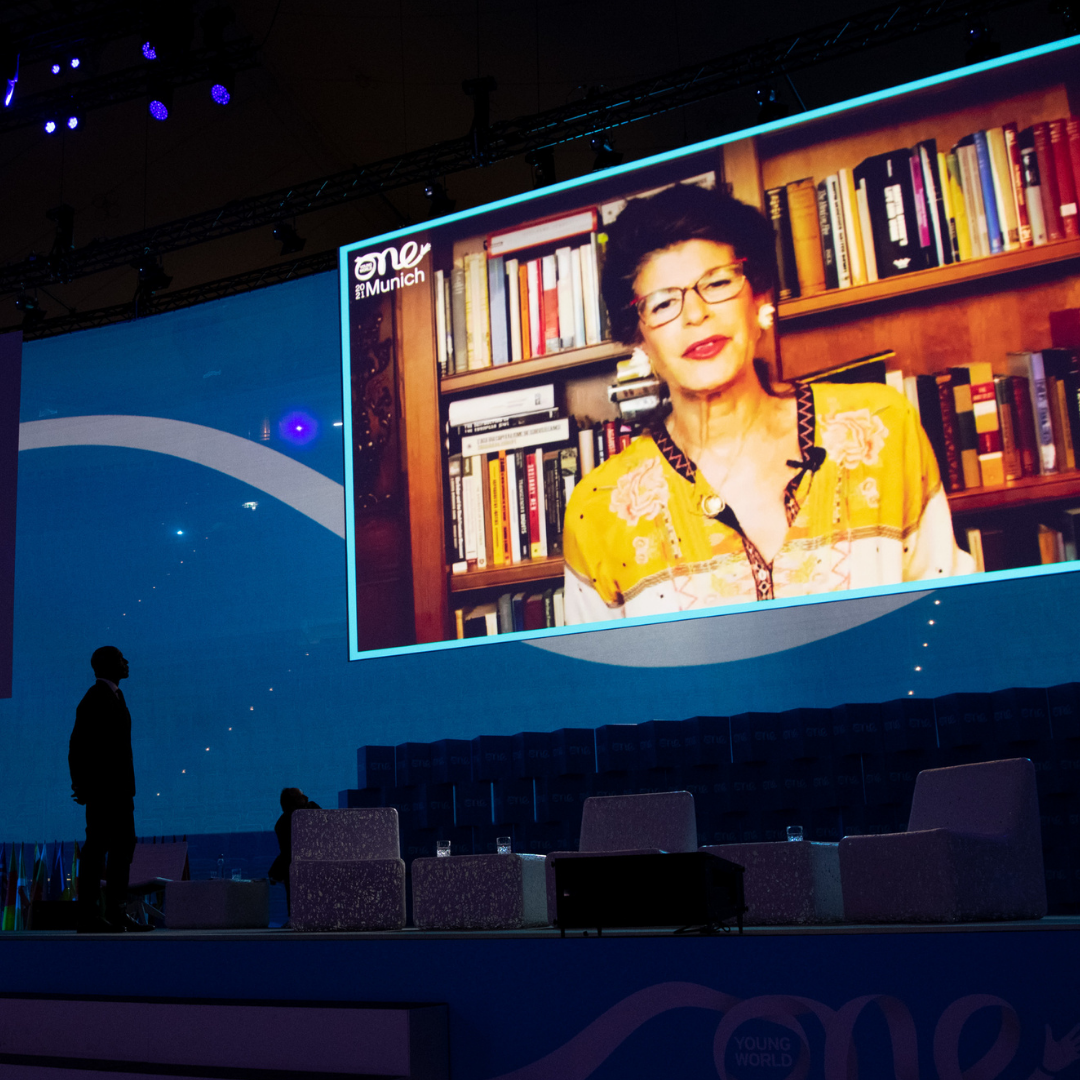 A screen is projected on a dark blue stage, a man can be seen standing looking up. One screen, a women speaks to the camera.