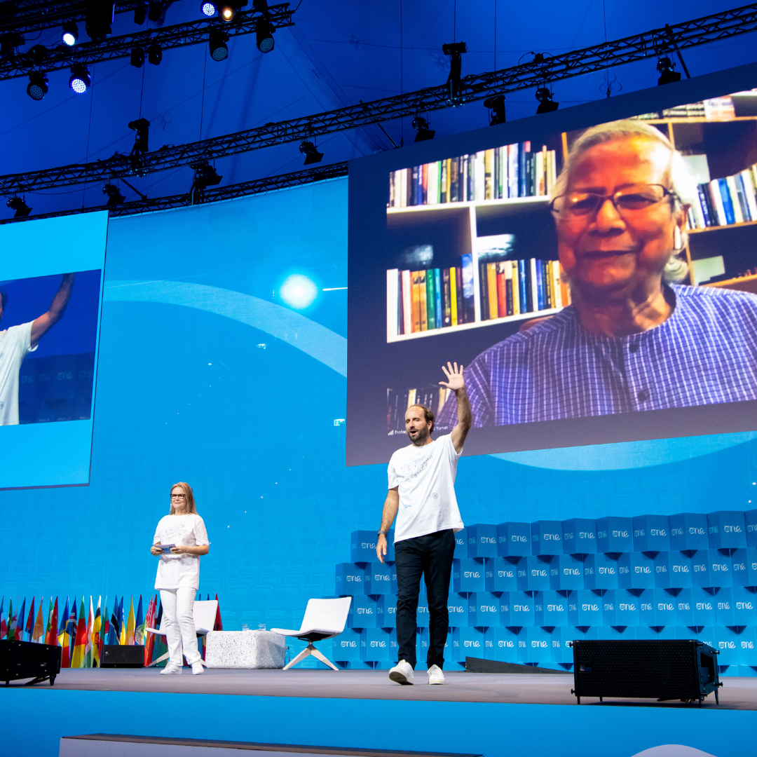 A man and a women stand on stage both wearing white t-shirts. She is in the background, he walks towards the front of the stage waving. A large screen is projected behind them featuring another man speaking to the camera.