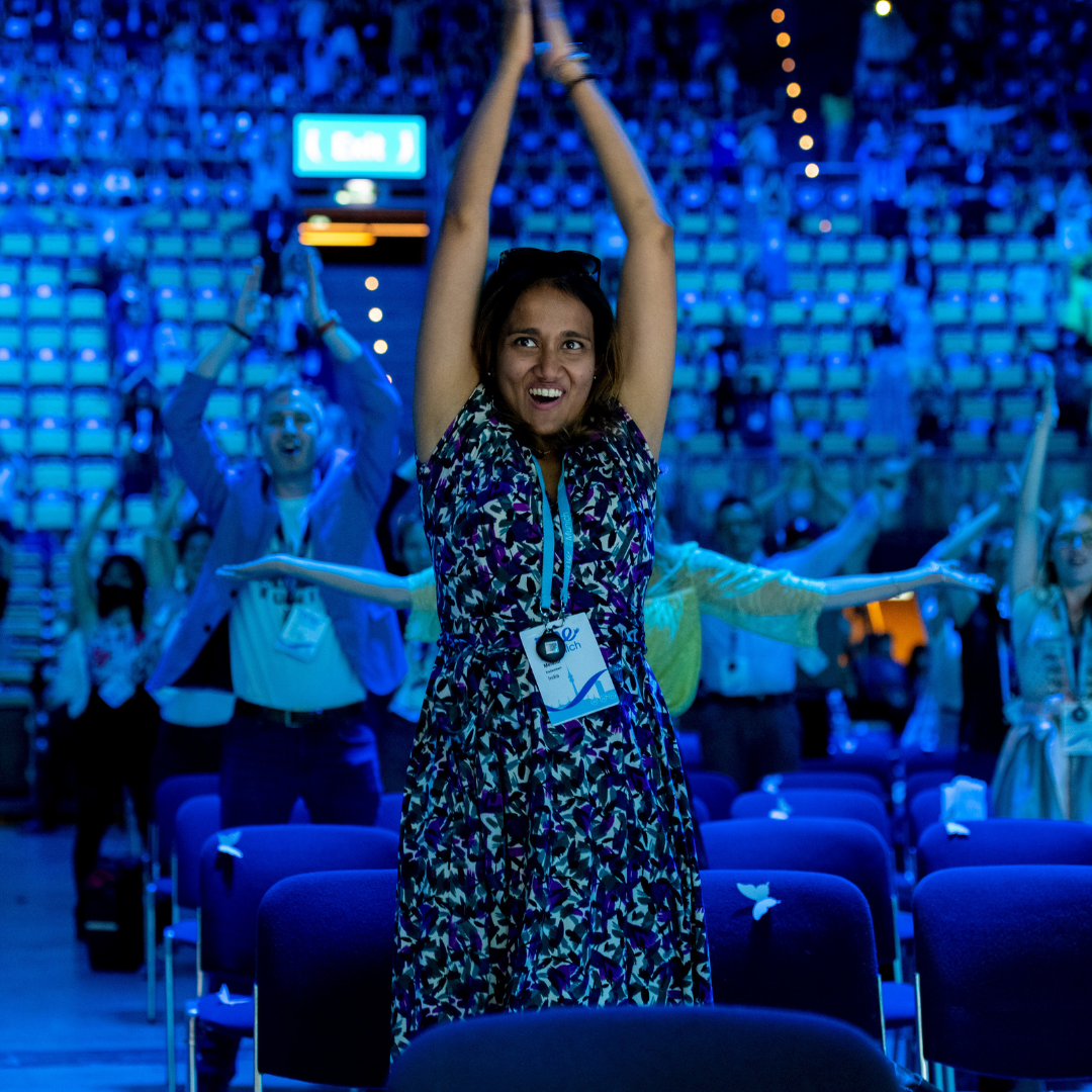 A young female Delegate claps her hands above her head smiling in the audience of an auditorium. Other members also mid-dance move can be seen in the background.
