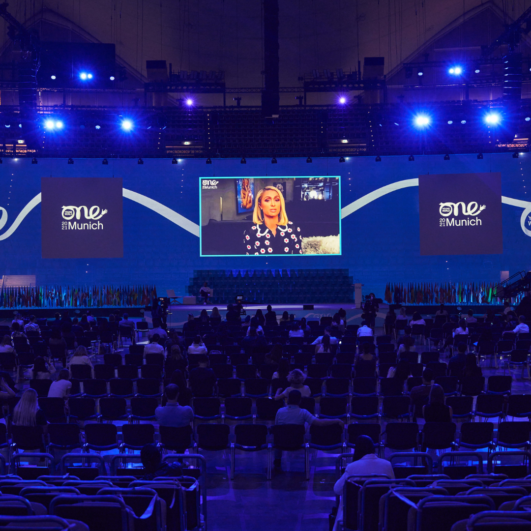 Taken from the back of the auditorium, a socially distanced audience sit basked in deep blue lighting. They are watching a women speak [Paris Hilton], projected on a large screen above a stage.