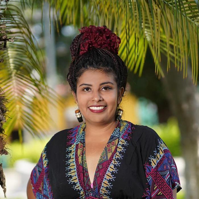 Ana Yency Photo wearing tribal pattern top, maroon head garment, set against tropical background