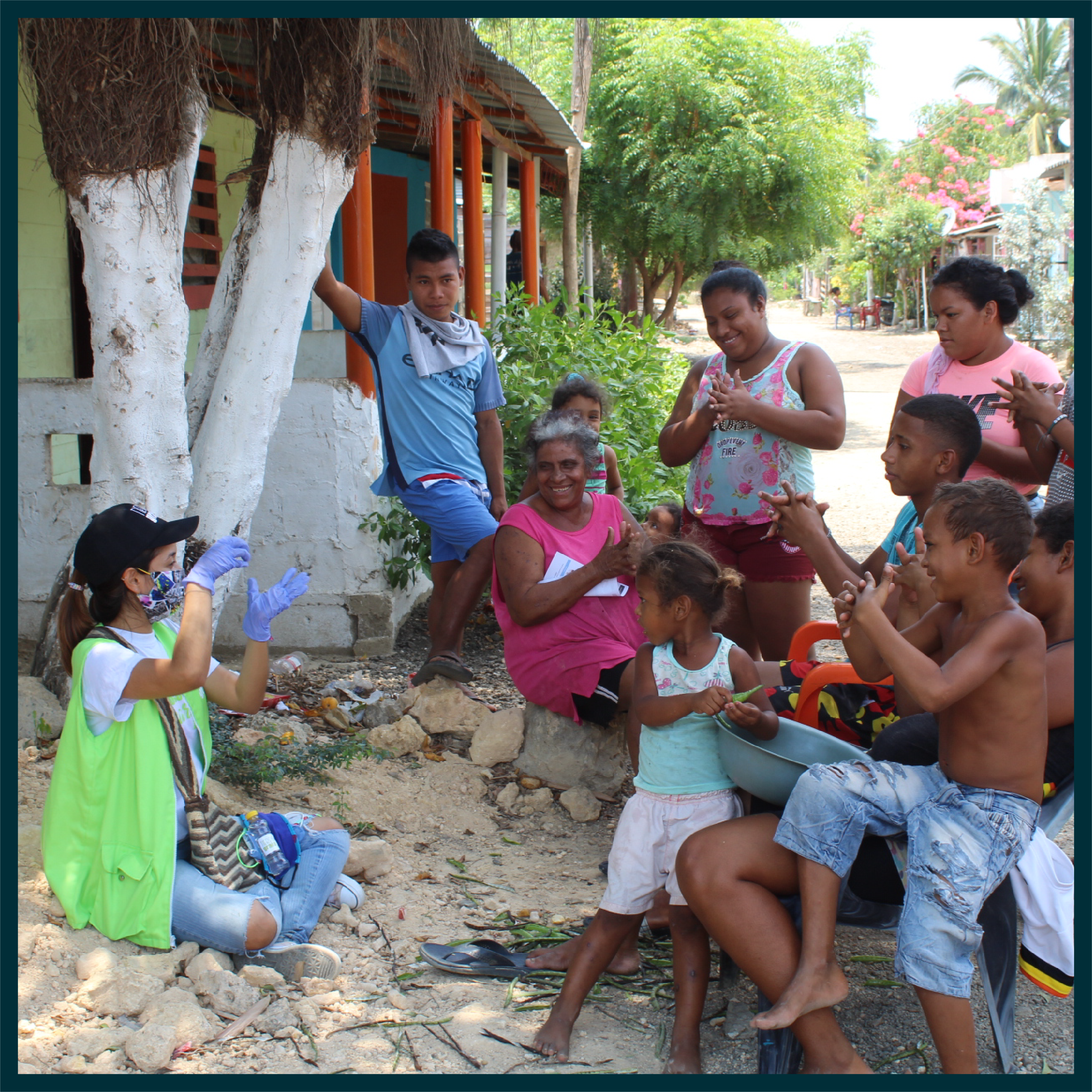 Image of One Young World Ambassador wearing PPE equipment, sat on the floor, explaining to a group of community members practices to reduce the spread of Covid-19