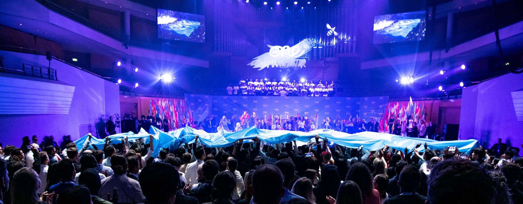 OYW banner passing over delegate audience crowd in front of the presentation stage at Manchester 2022 Summit