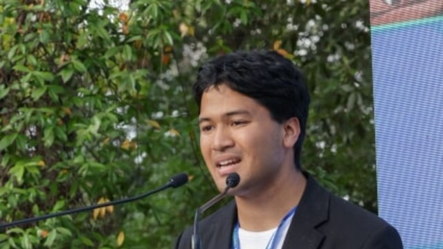 Hikaru presenting at a podium against green hedge background