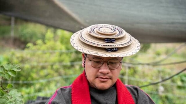 Leider Andres Tombe Morales portant un chapeau rond et orné, un vêtement de cou rouge parmi des arbustes à hauteur de la taille et des épaules.