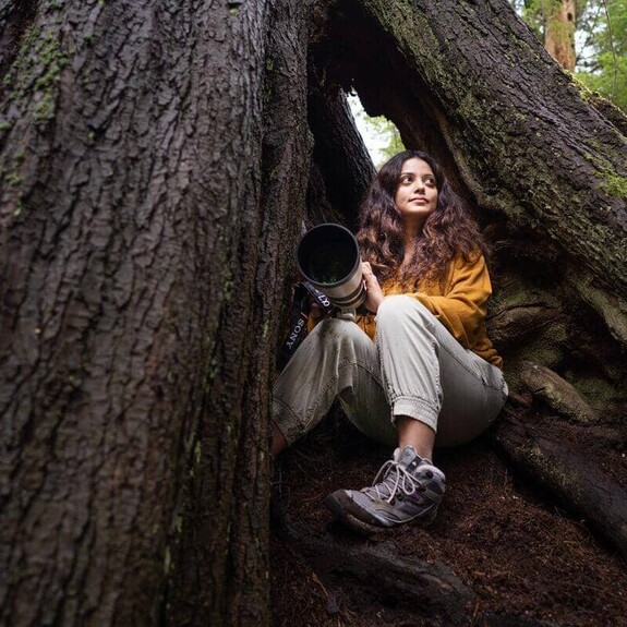 Gunjan Menon sitting at the base of a tree facing up holding a digital camera