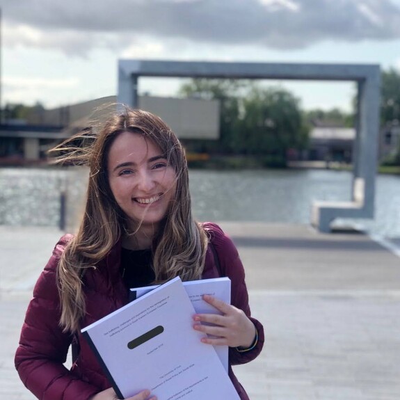 Anxhela Bruci wearing dark dark pink gillet holding documents on a riverside front