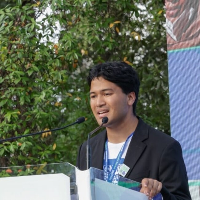 Hikaru presenting at a podium against green hedge background