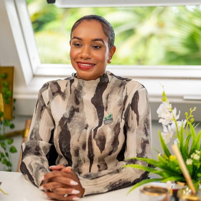 Keithlin Caroo-Afrifa in pattern top sitting at desk surrounded by green indoor plants