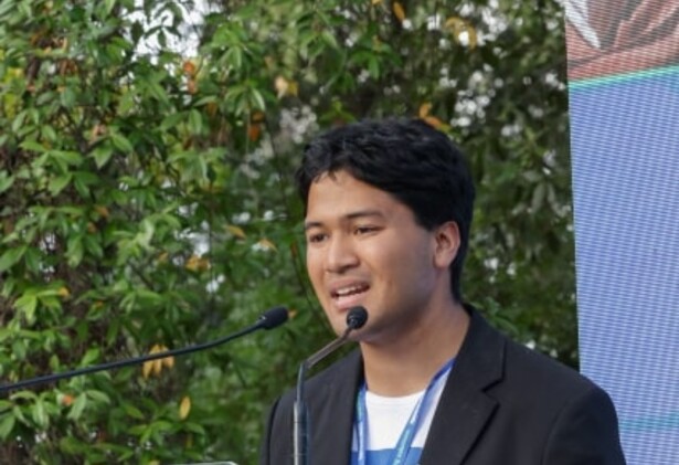 Hikaru presenting at a podium against green hedge background