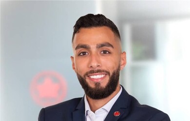 Abdullah El-Safadi portrait, wearing dark blue navy blazer with red pin on lapel, white shirt against grey blurred background