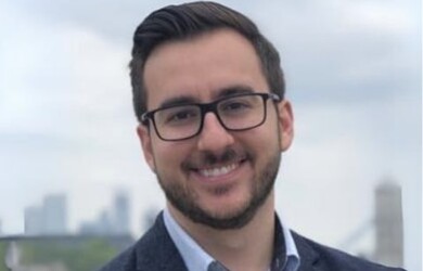 Bradley portrait, wearing navy blazer and blue shirt against cityscape background