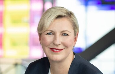 Emmanuelle portrait wearing white undershirt dark blue jacket and multi-coloured lapel pin against neon lights background