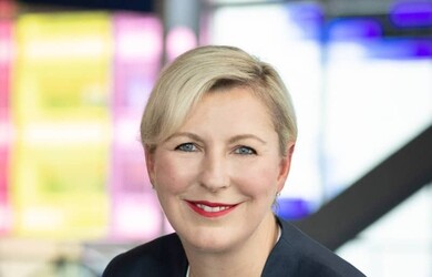 Emmanuelle portrait wearing white undershirt dark blue jacket and multi-coloured lapel pin against neon lights background