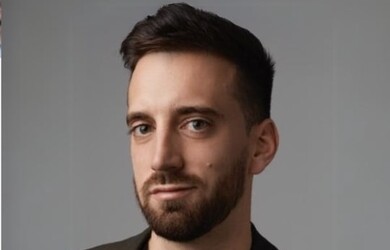 Simon portrait wearing black undershirt and brown blazer against grey background