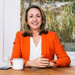 Dr. Patricia Widmer wearing white undershirt and orange blazer sitting at a table with grass painting in the background