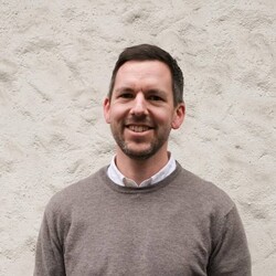 Florian Hoffmann portrait wearing shirt and crew neck light brown jumper against textured beige background